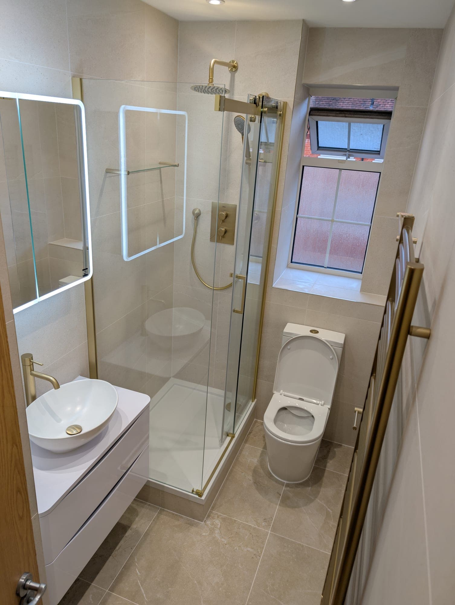 Corner shower with a brushed brass frame and rainfall head, round countertop basin on a white floating vanity, backlit mirror cabinet and sash window with obscured glazing