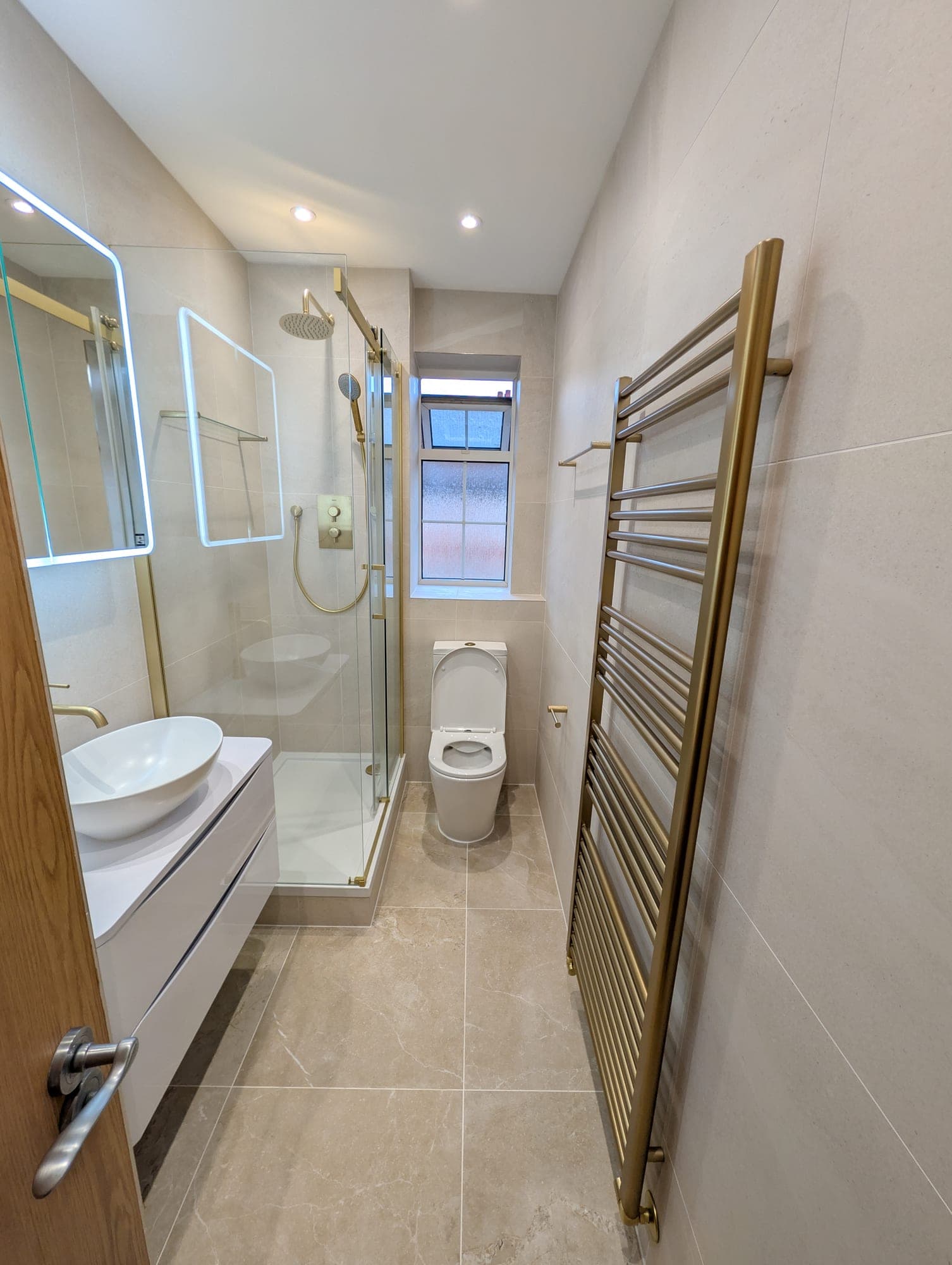 Brushed brass heated towel rail beside a corner shower with rainfall head, round countertop basin and floating white vanity reflected in the backlit mirror