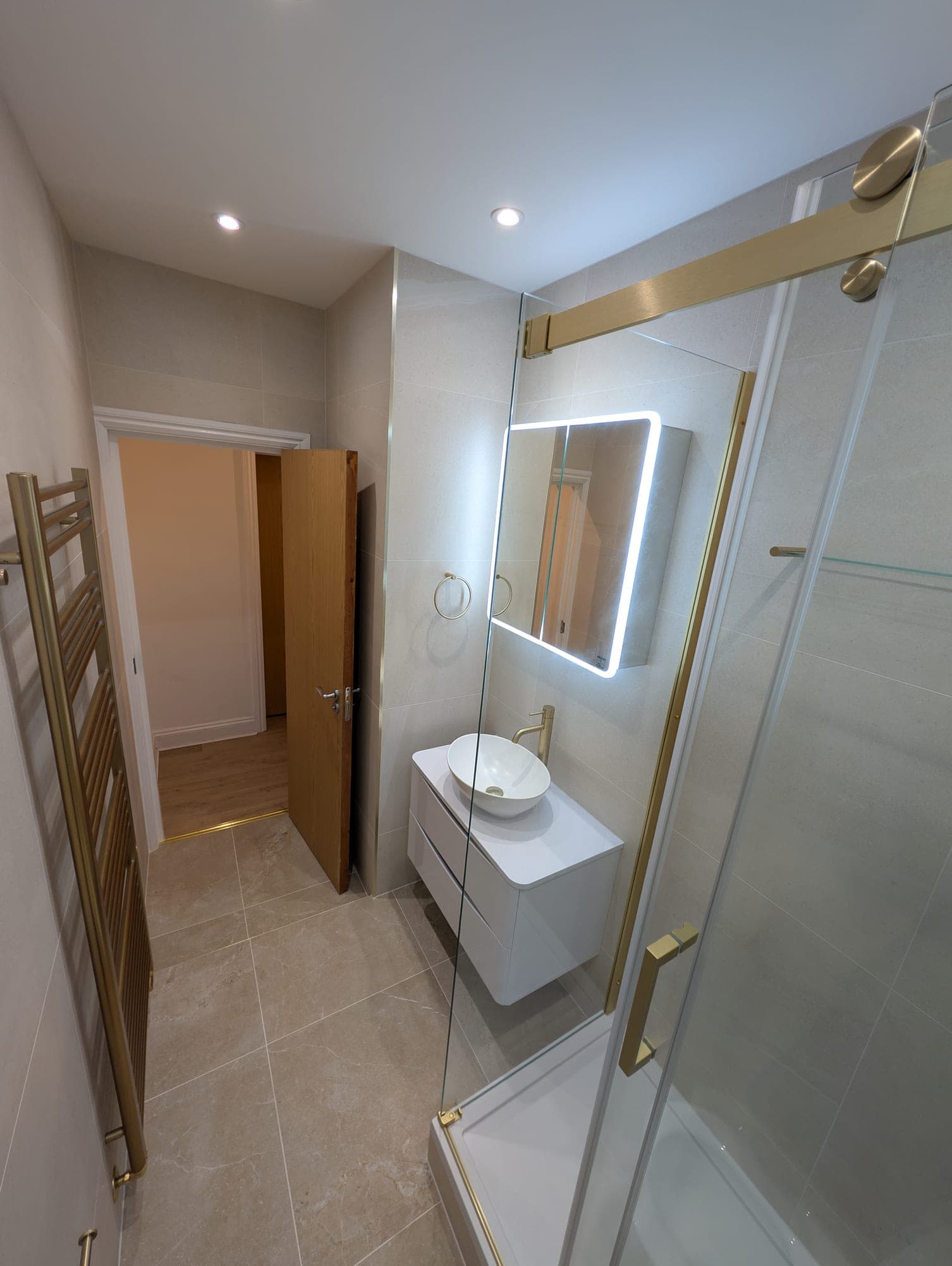 Round countertop basin on a white floating vanity with brushed brass tap, backlit mirror cabinet and an open door onto a hallway with wood flooring