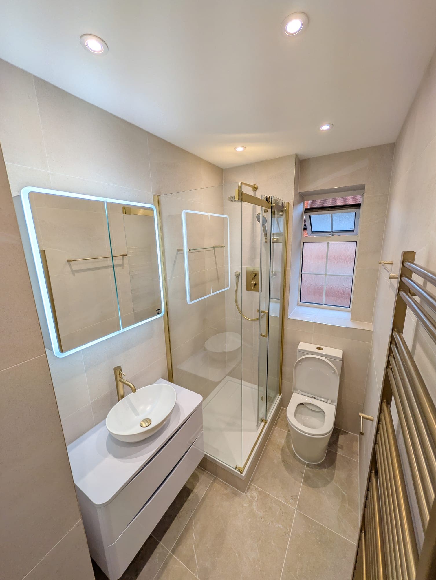 White floating vanity with round countertop basin and brushed brass fittings beside a brass-framed corner shower enclosure and obscured-glass window