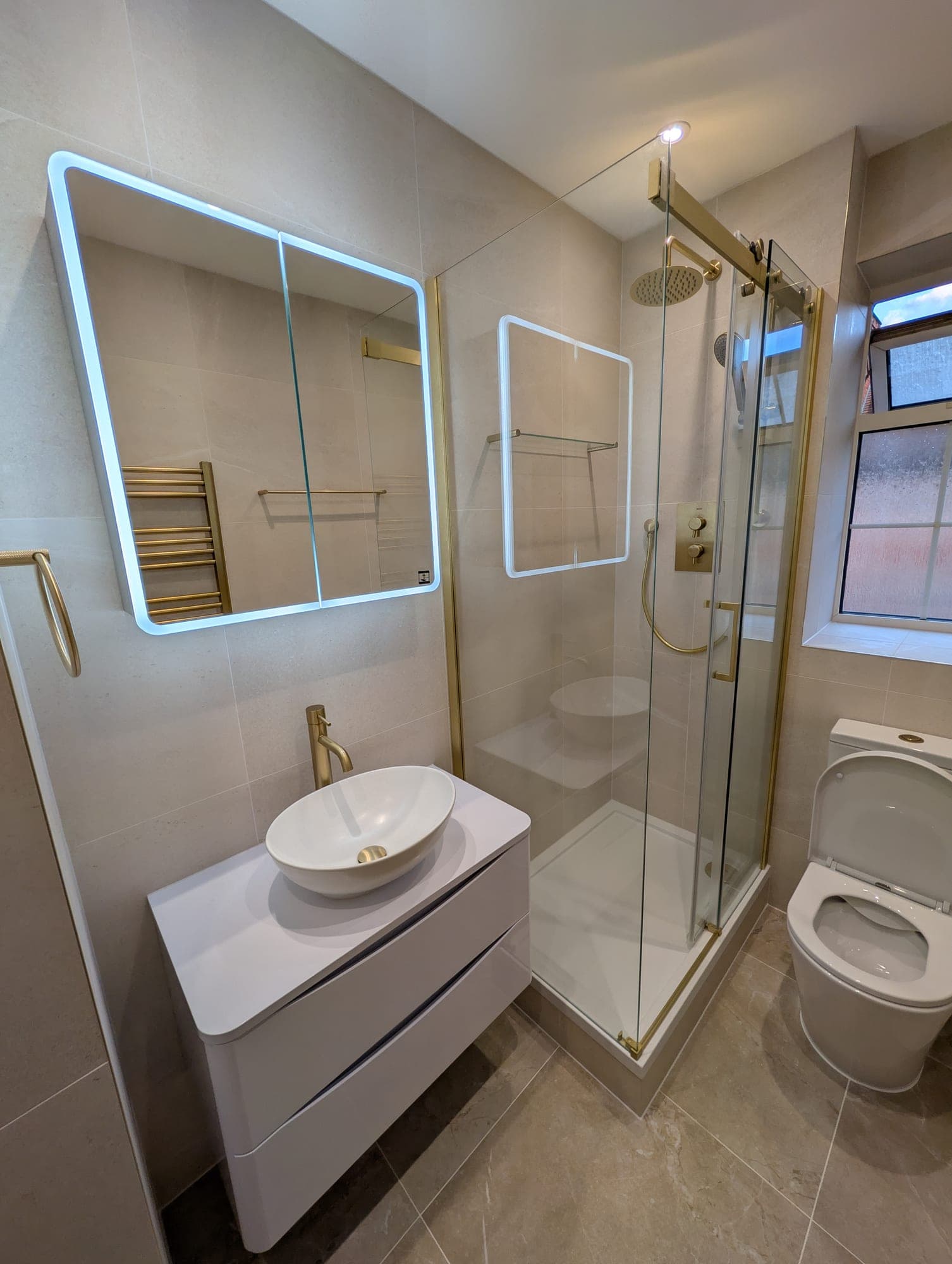 Overview of an en-suite with brass-framed corner shower, round countertop basin, backlit mirror cabinet and wall-hung toilet beneath an obscured-glass window
