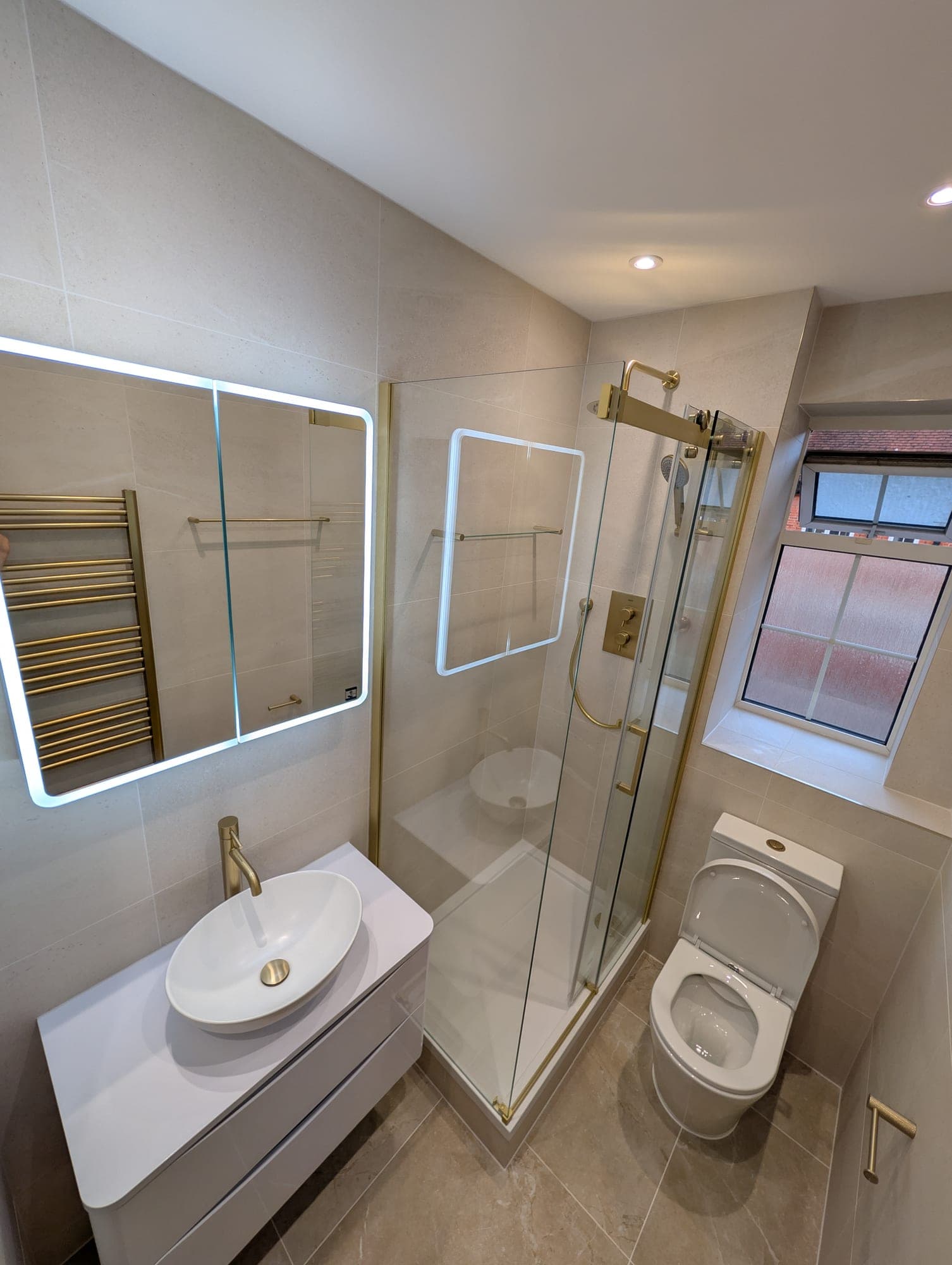Brass-framed corner shower and round countertop basin reflected in a backlit mirror, wall-hung toilet and heated towel rail alongside