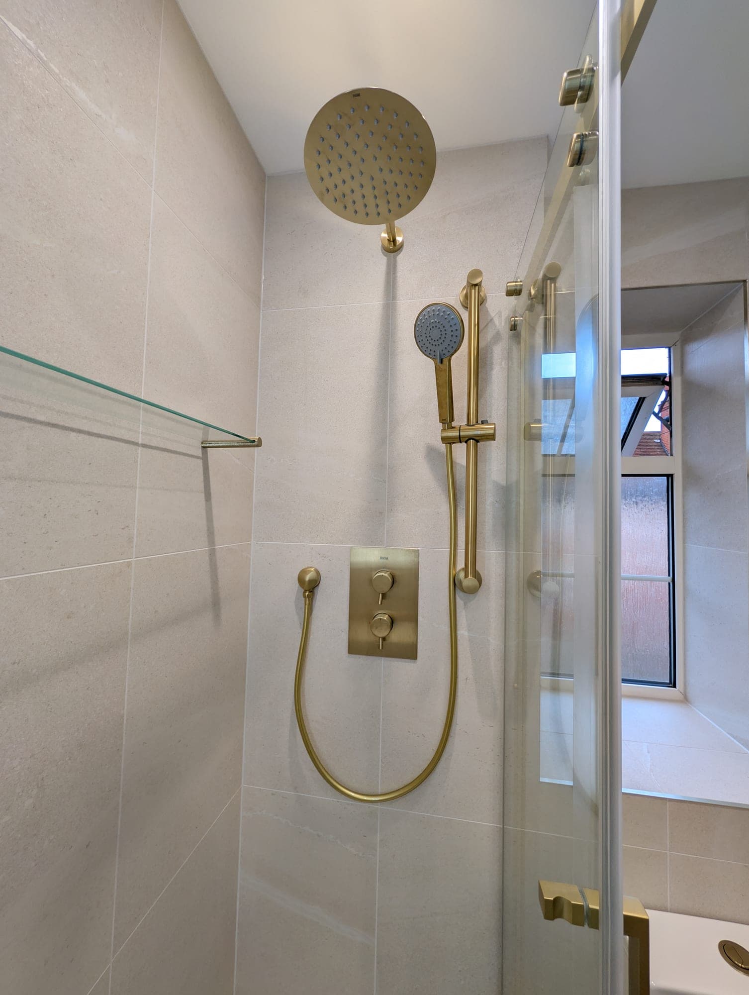 Close-up of a brushed brass shower riser rail, handset and rainfall head against stone-effect tiling in a brass-framed shower enclosure