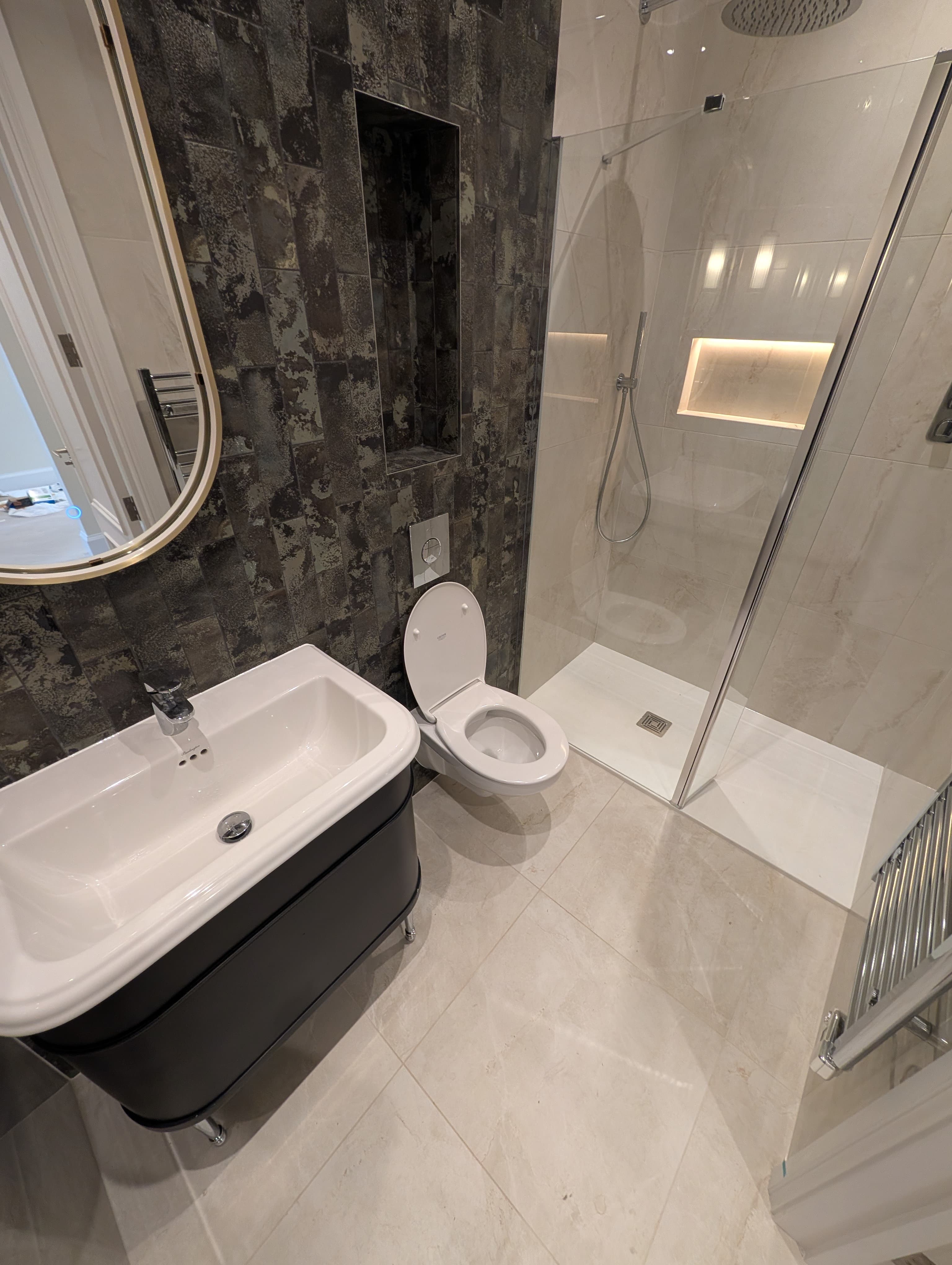 Dark textured tile feature wall with a recessed shower niche and wall-hung toilet, glass shower screen and brass mirror edge visible alongside