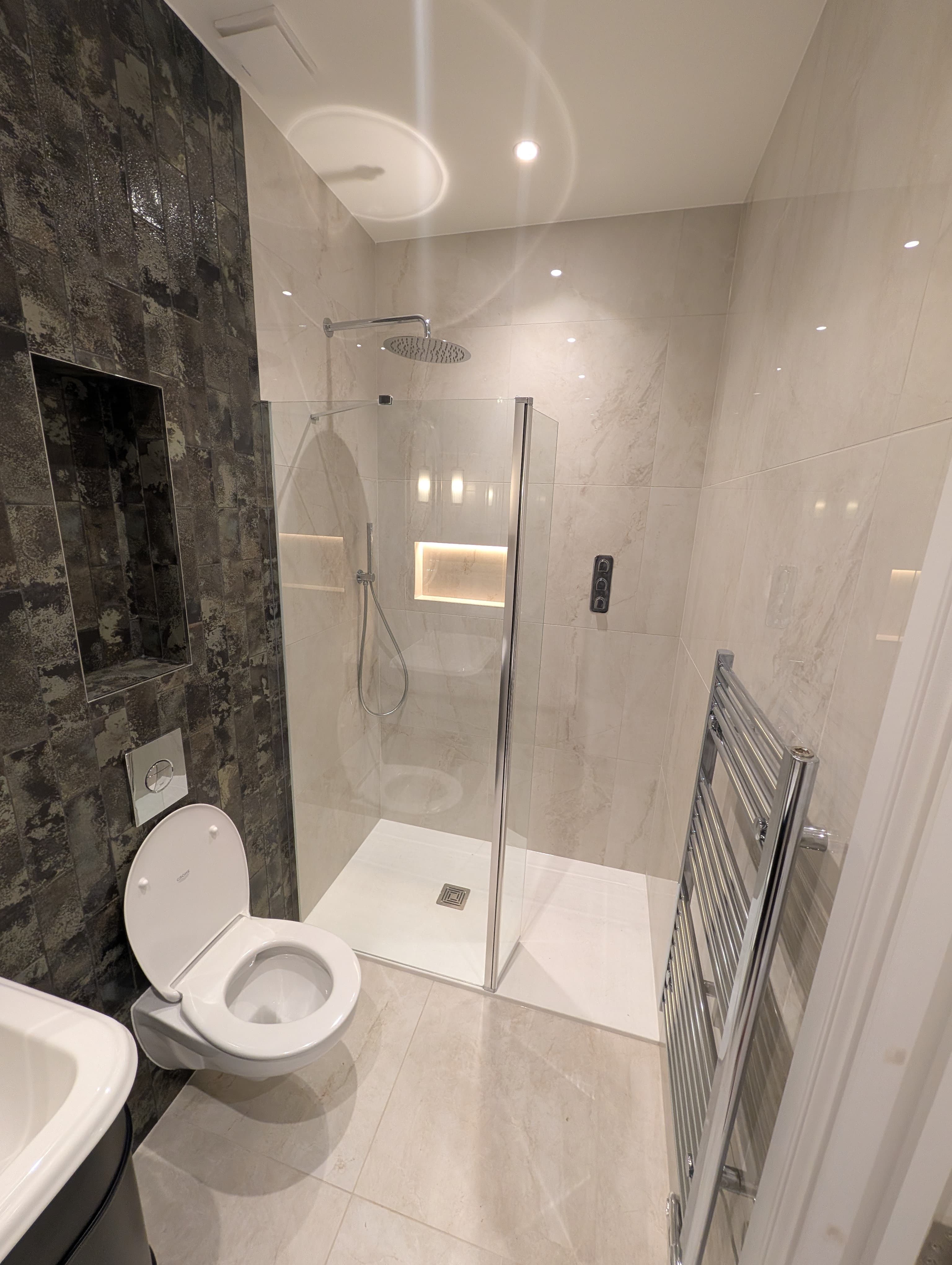 Walk-in glass shower with ceiling-mounted rainfall head and pale stone-effect tiling, chrome heated towel rail alongside