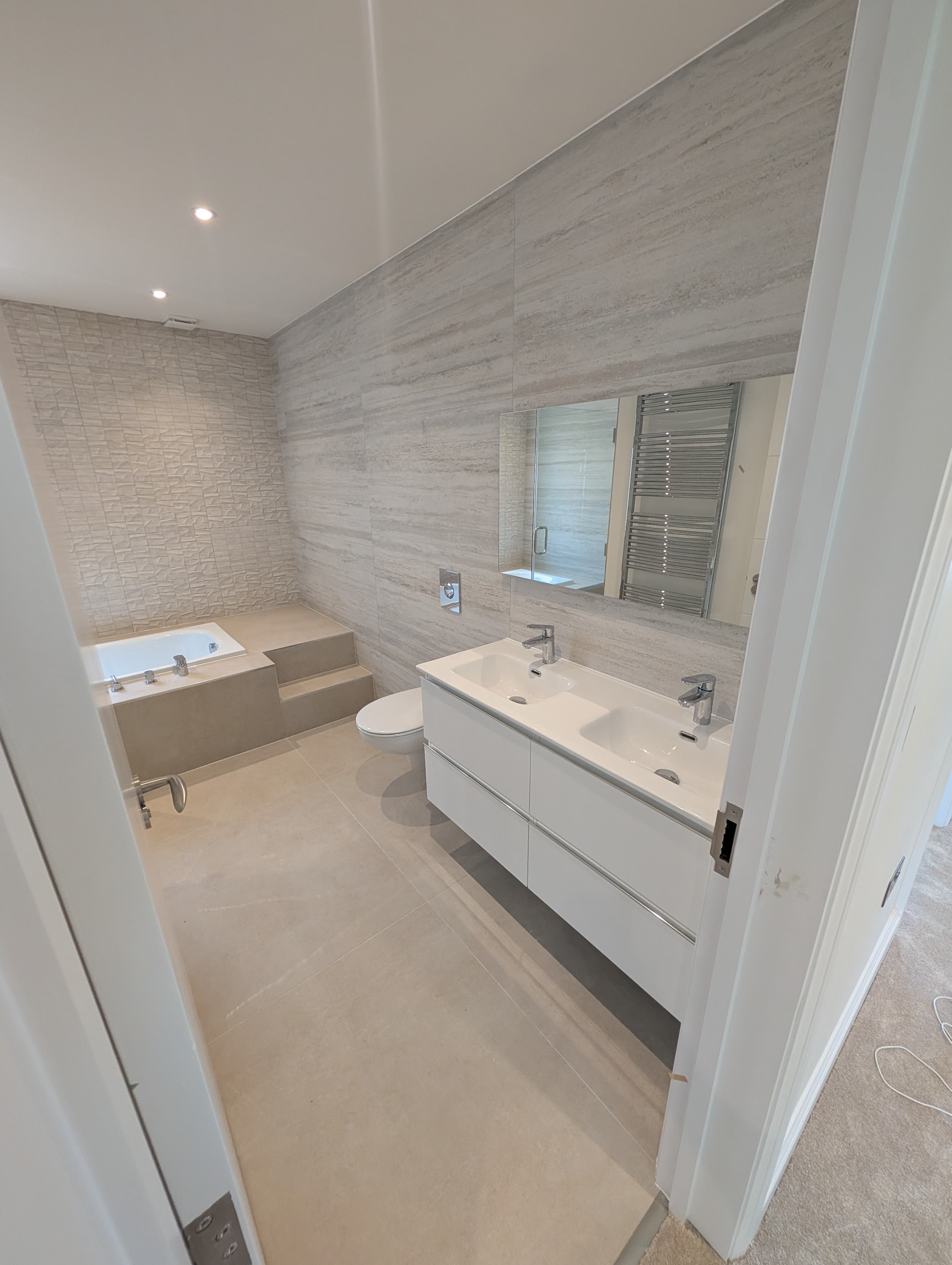 Double vanity with white countertop basins and chrome taps beneath a large mirror reflecting a chrome heated towel rail, raised bath visible alongside