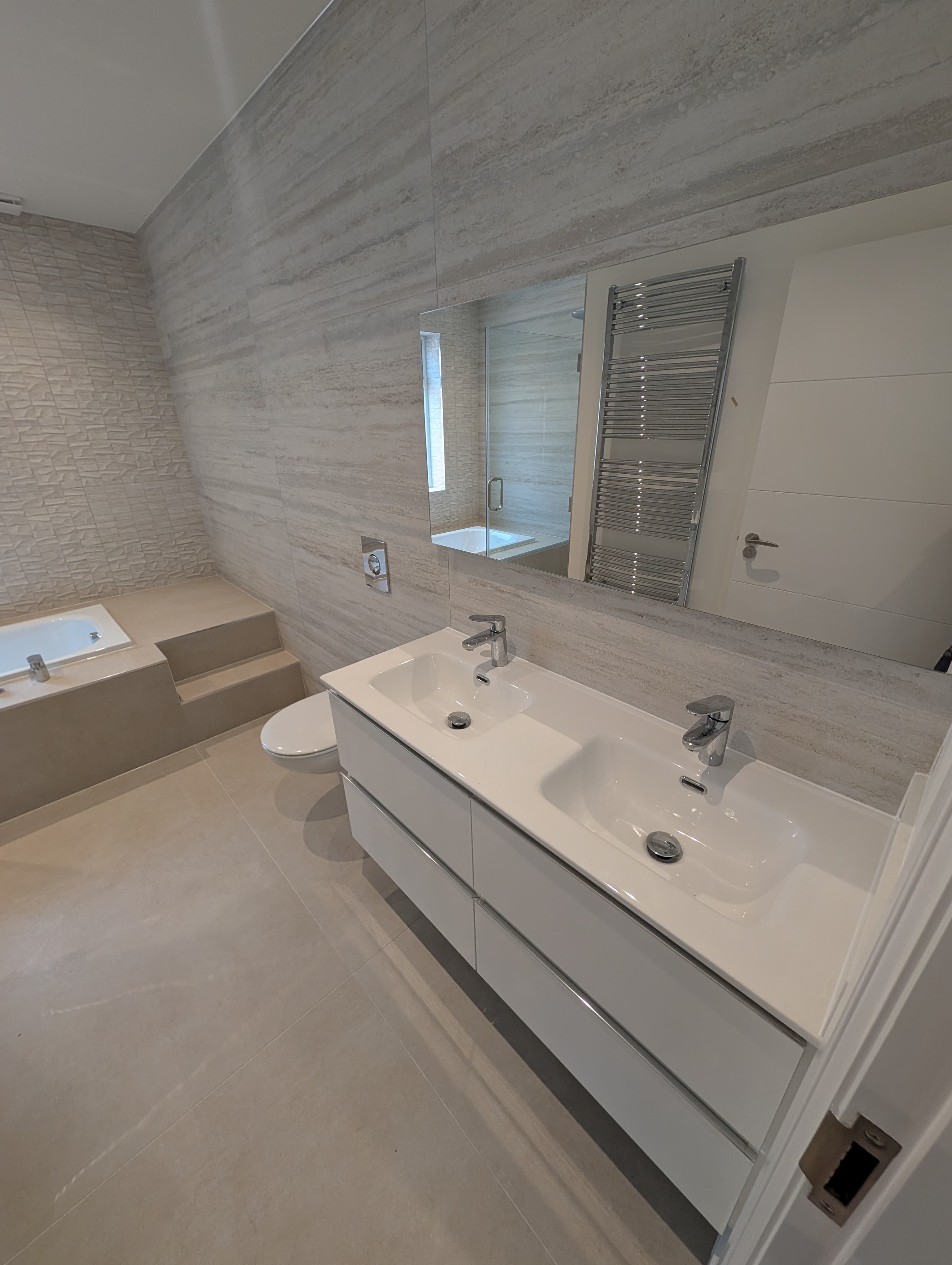 Bath on a raised tiled step below a window, glass shower screen and textured tile feature wall alongside