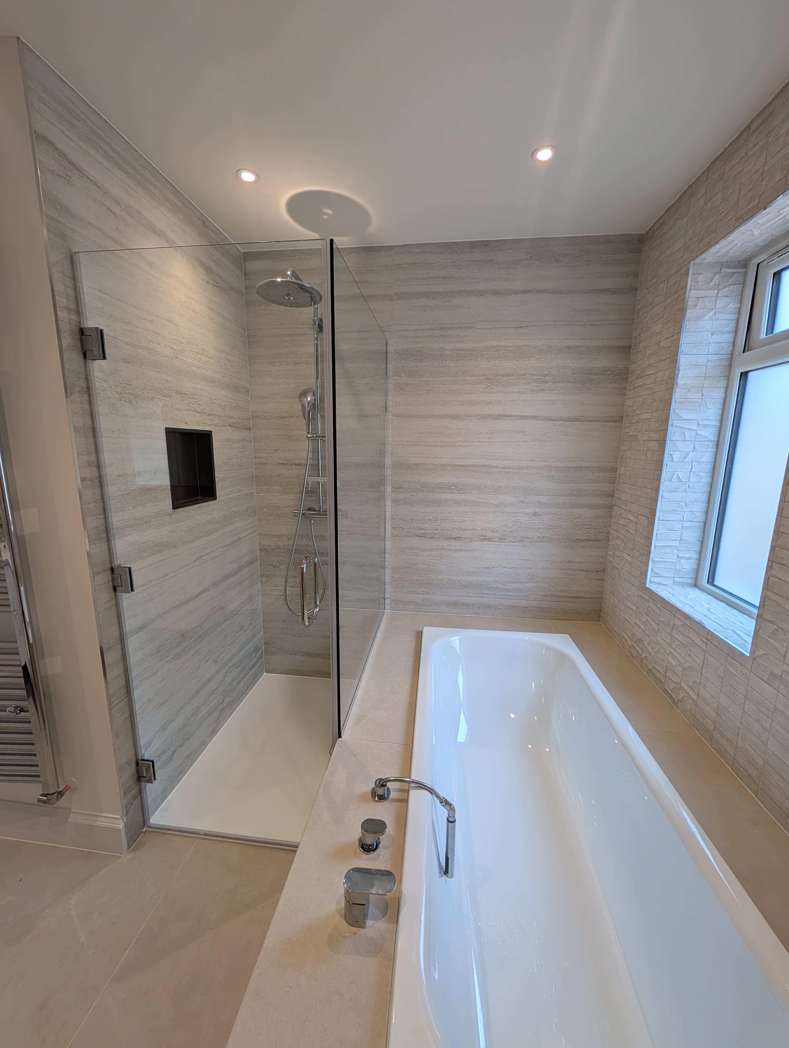 Glass-screened walk-in shower with rainfall head and recessed niche beside a raised bath and window in a textured tile en-suite