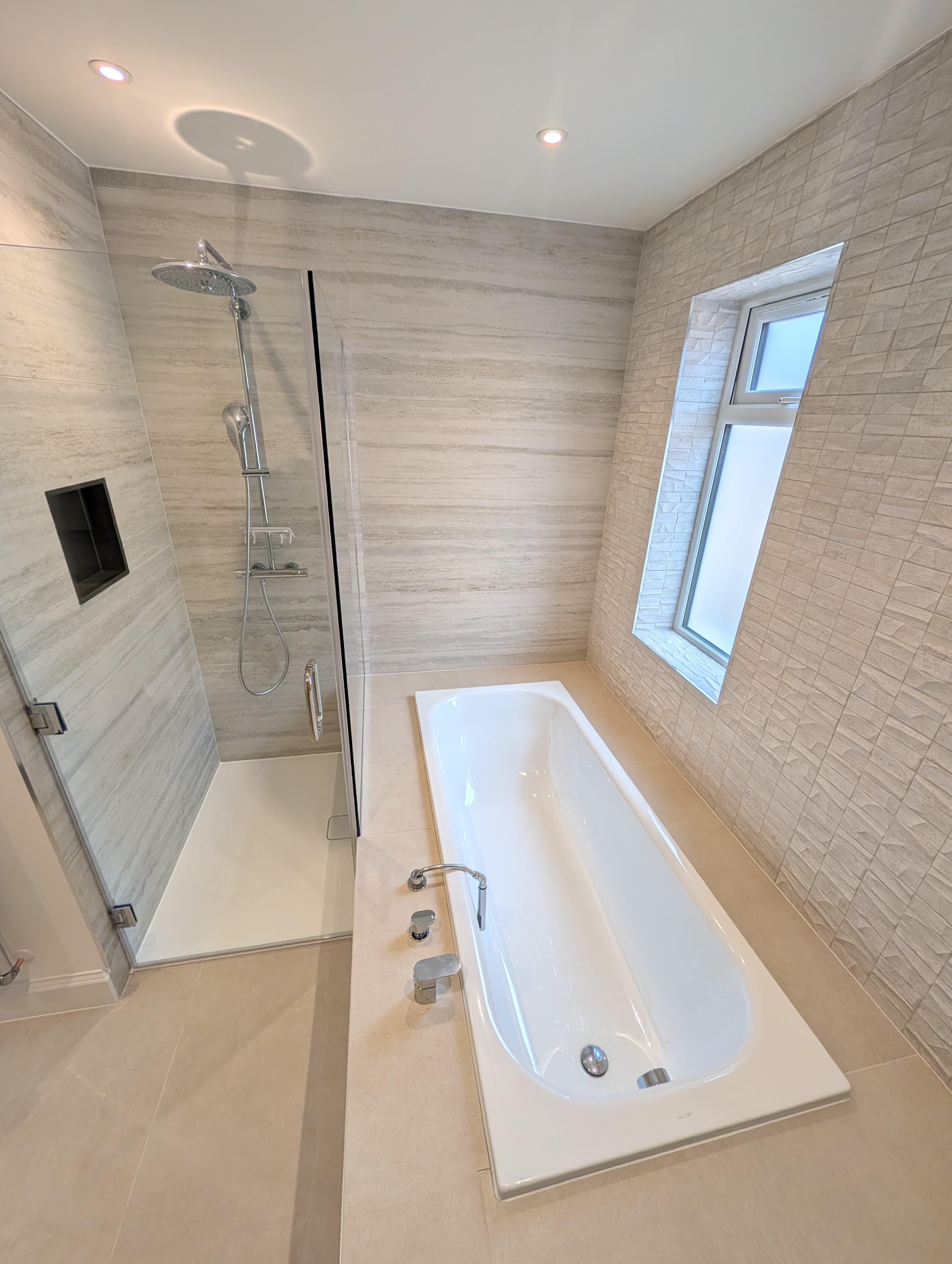 Walk-in shower with rainfall head and recessed niche beside a raised bath below a textured tile feature wall and window