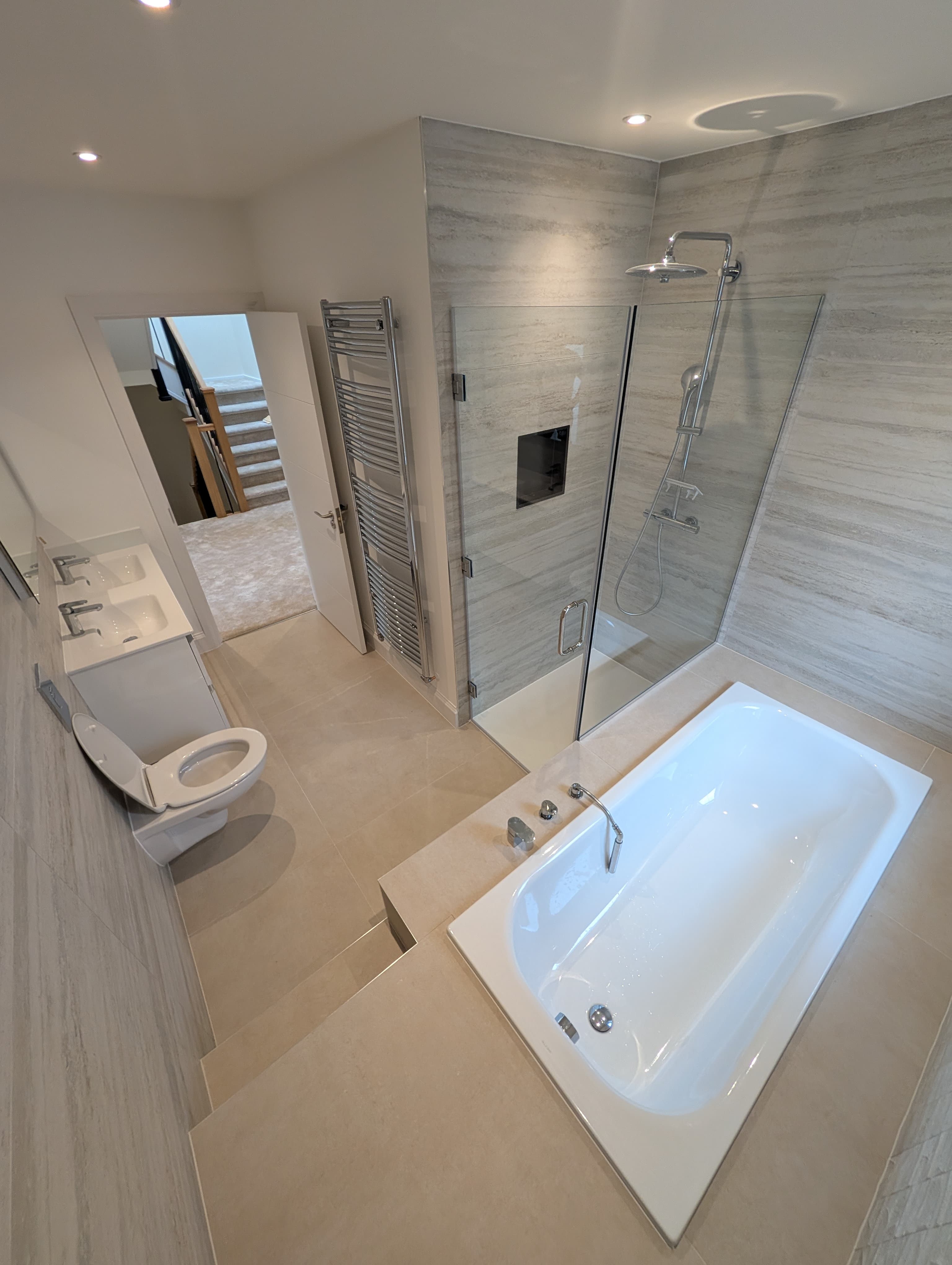 Overview of an en-suite with glass shower, raised bath and wall-hung toilet, chrome heated towel rail and doorway to the landing