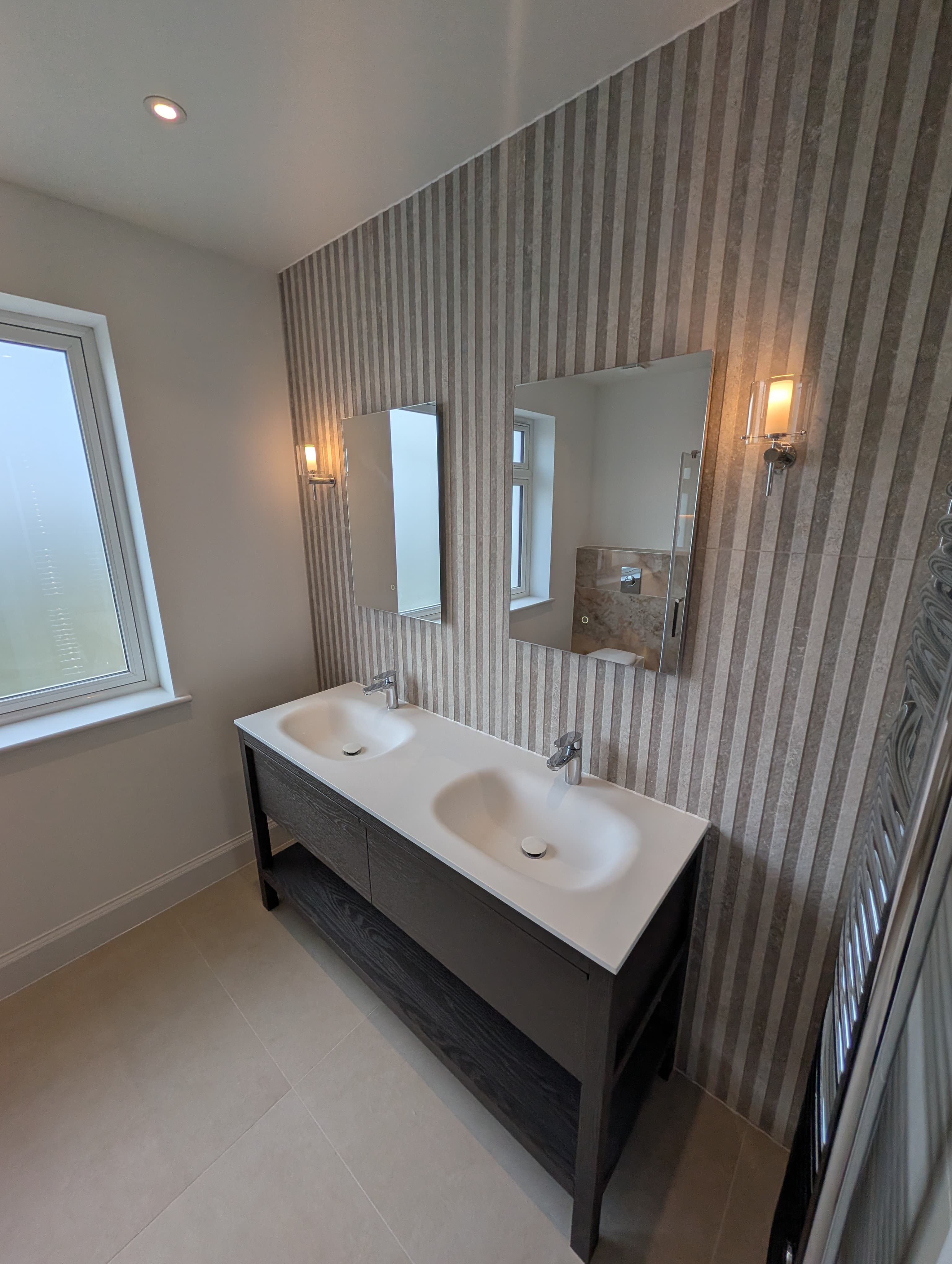 Double vanity in dark stained wood with white countertop basins, twin mirrors and a striped wallpaper feature wall, chrome heated towel rail alongside
