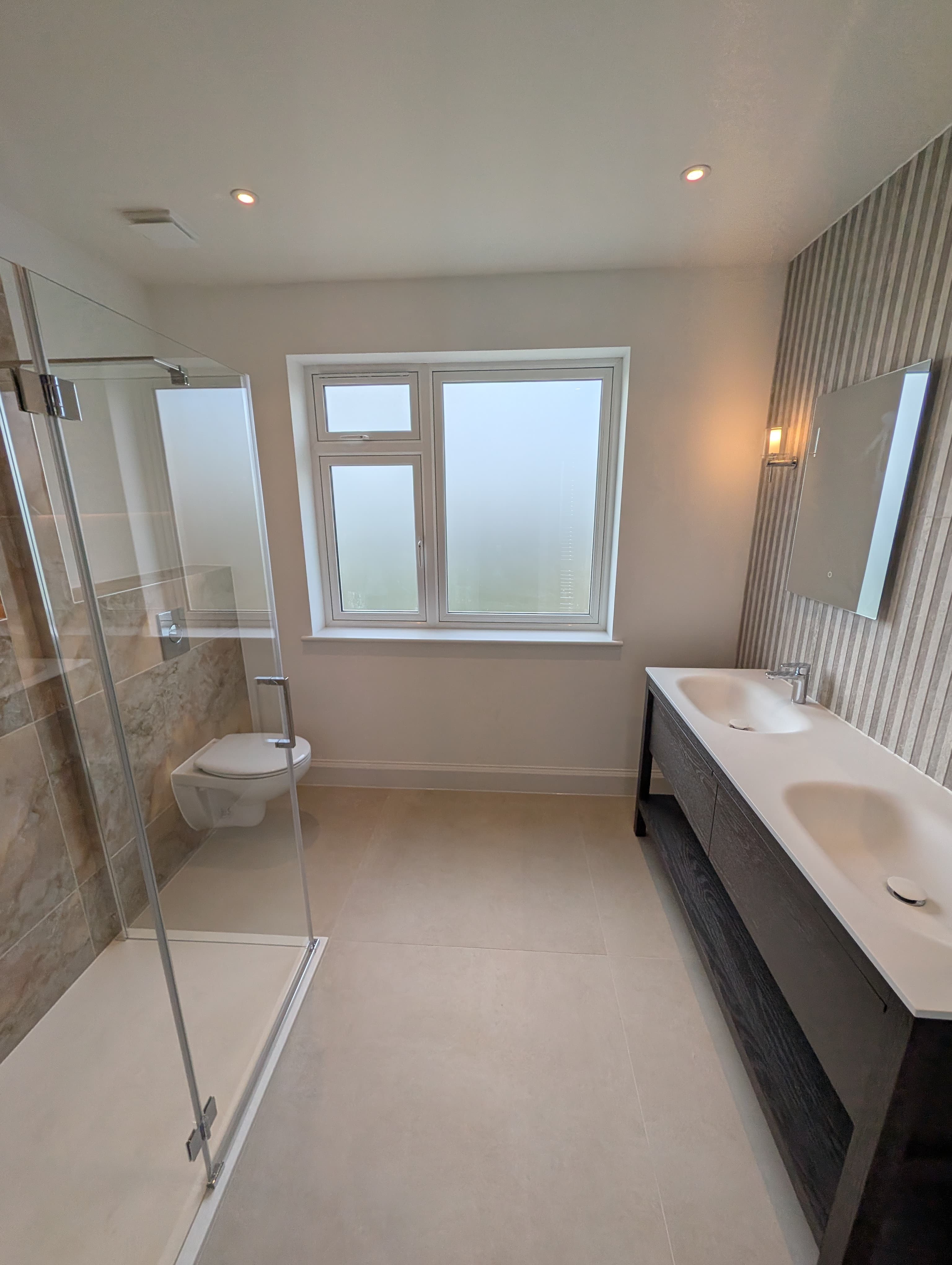 Corner glass shower and wall-hung toilet beside a tall window, dark stained wood double vanity visible in the background