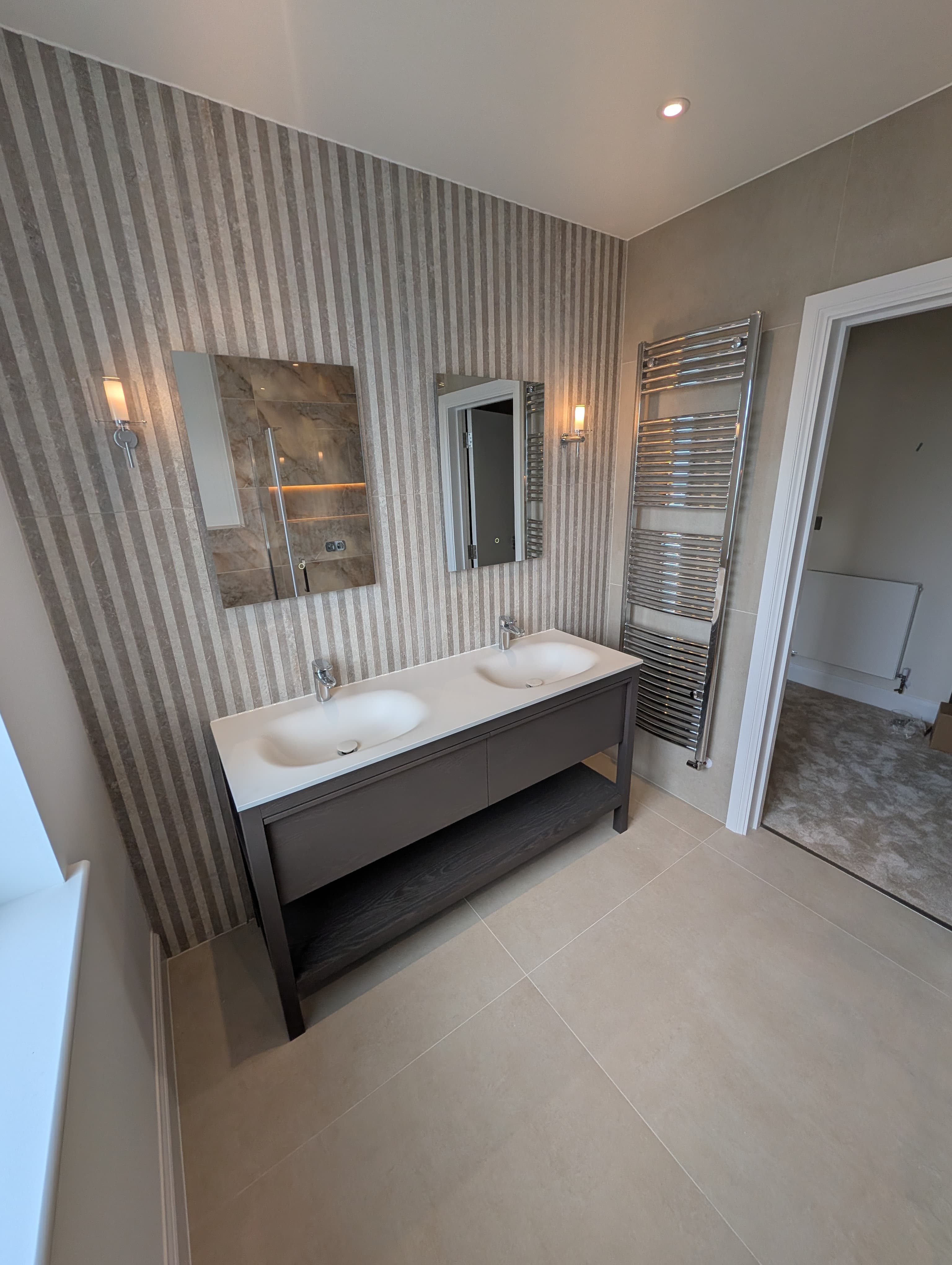 Dark stained wood double vanity with white countertop basins beneath twin mirrors on a striped wallpaper wall, chrome heated towel rail alongside