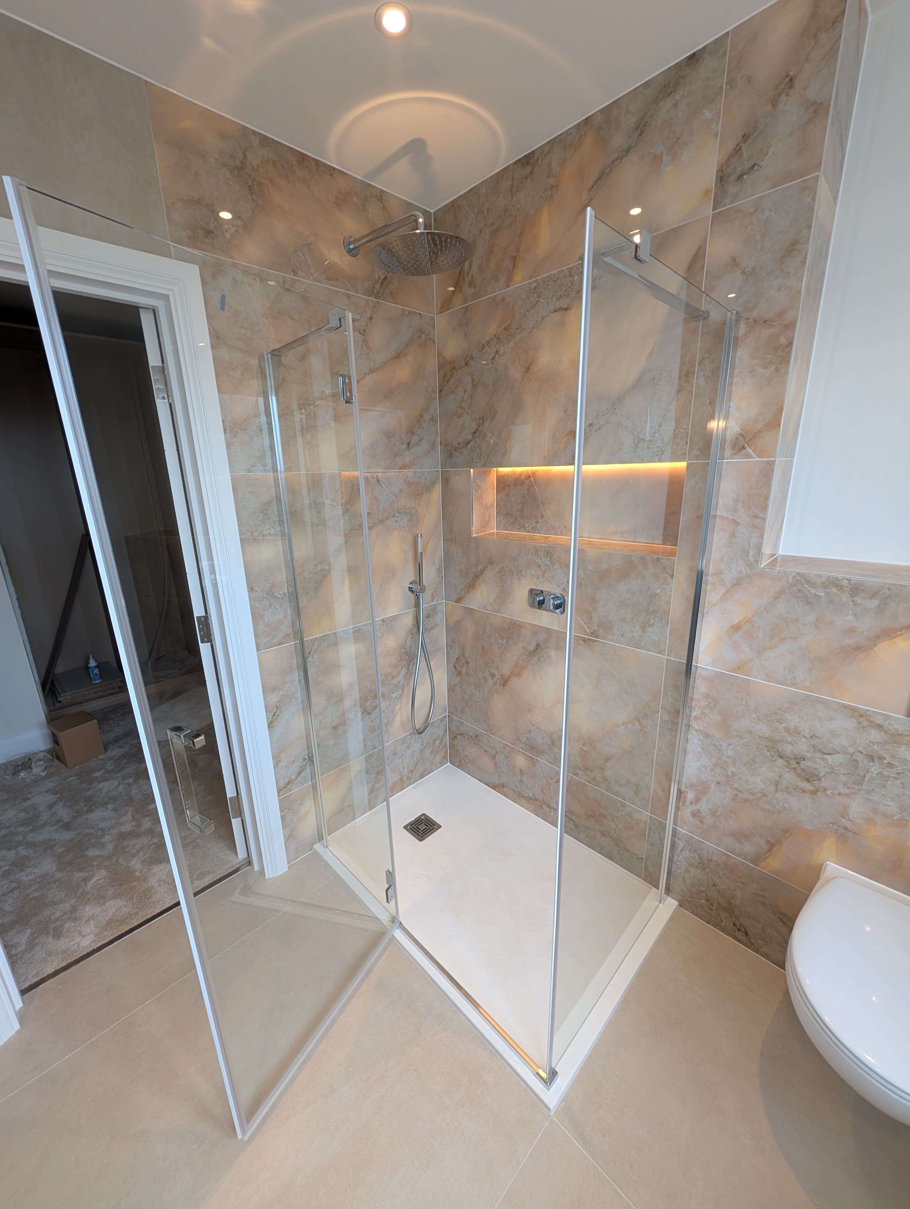 Frameless glass corner shower with warm marble-effect tiling, a backlit niche and ceiling-mounted rainfall head