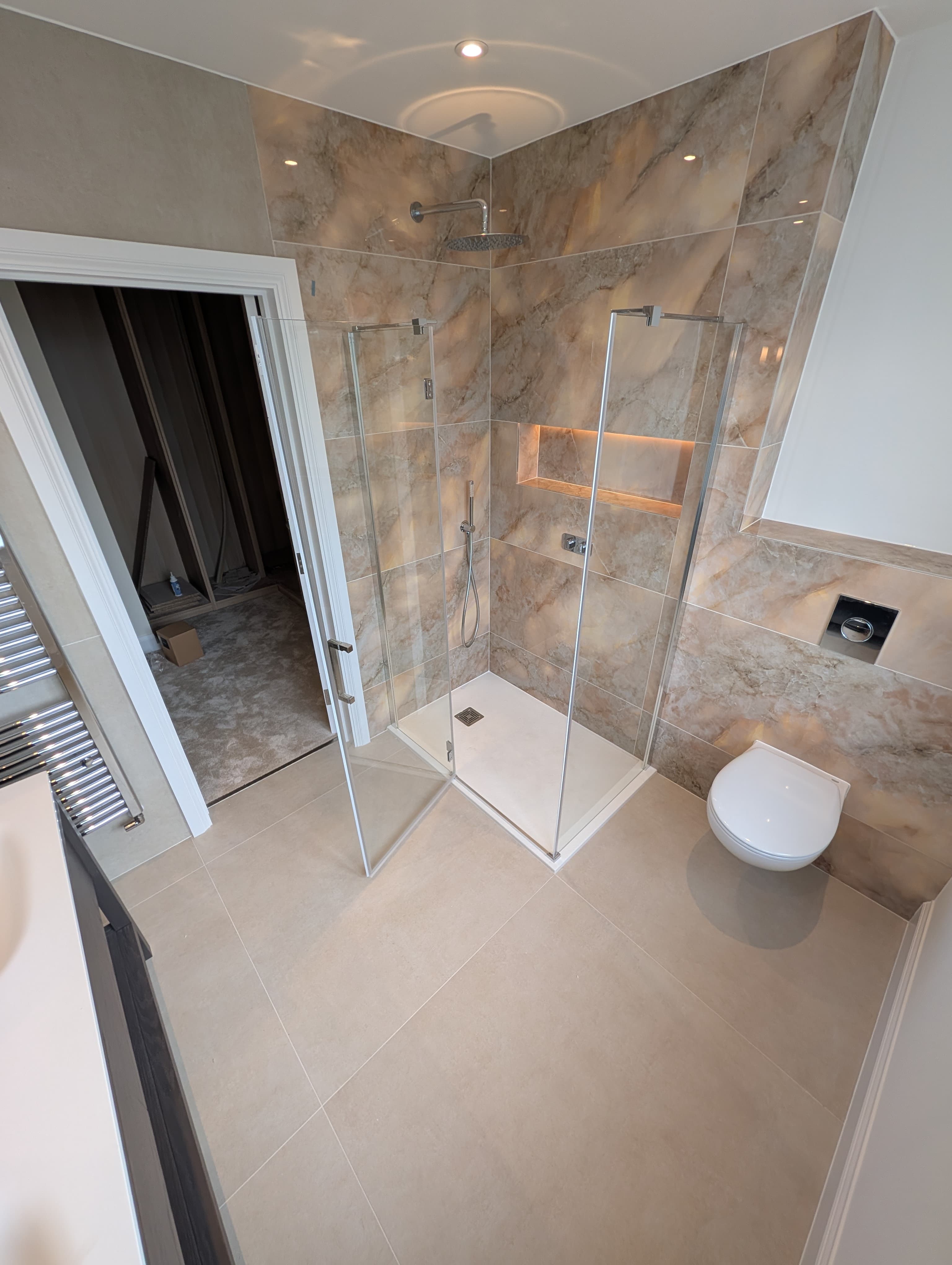 Marble-effect corner shower with a backlit recessed niche, chrome rainfall head and a wall-hung toilet alongside