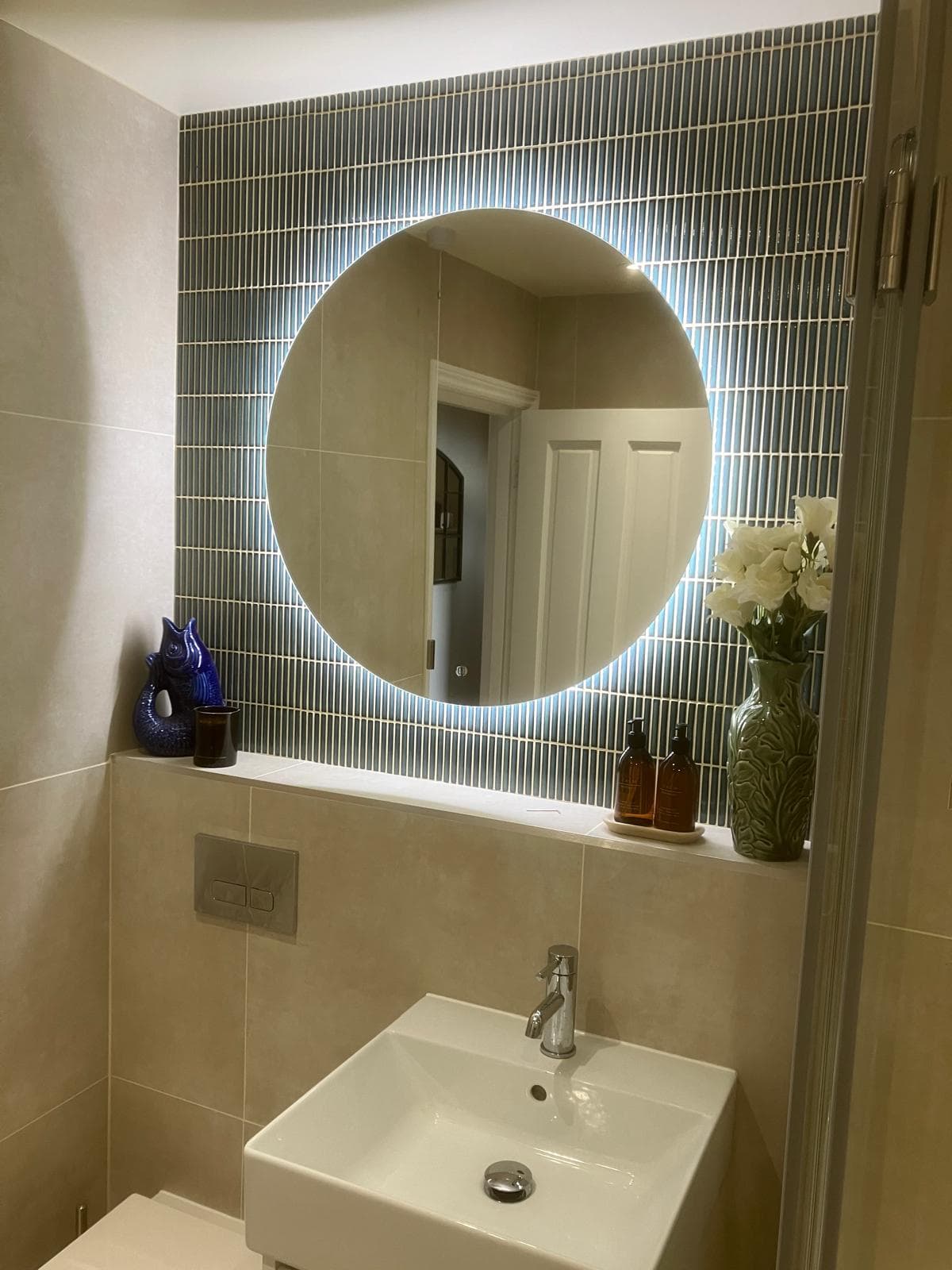 Round backlit mirror over teal subway-tiled splashback with wall-mounted sink