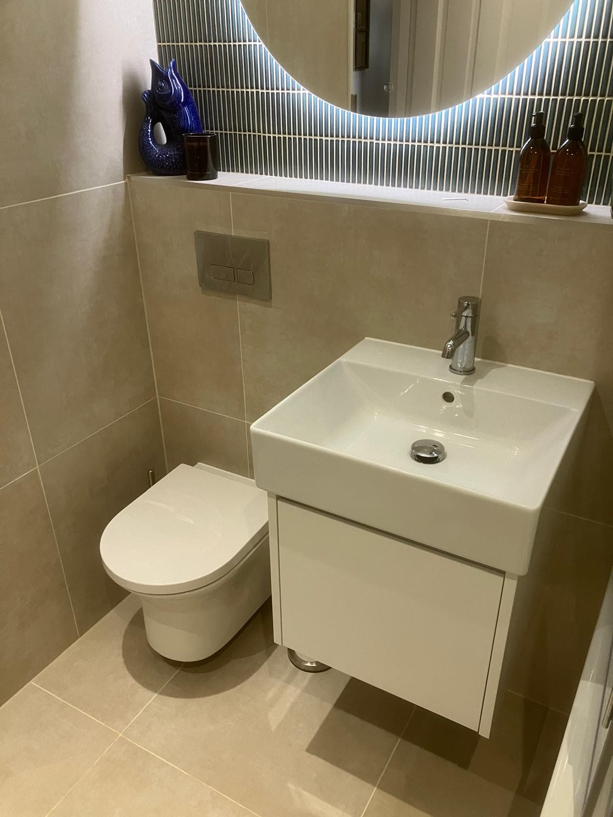 Sink and toilet beneath round backlit mirror with blue accent tile wall