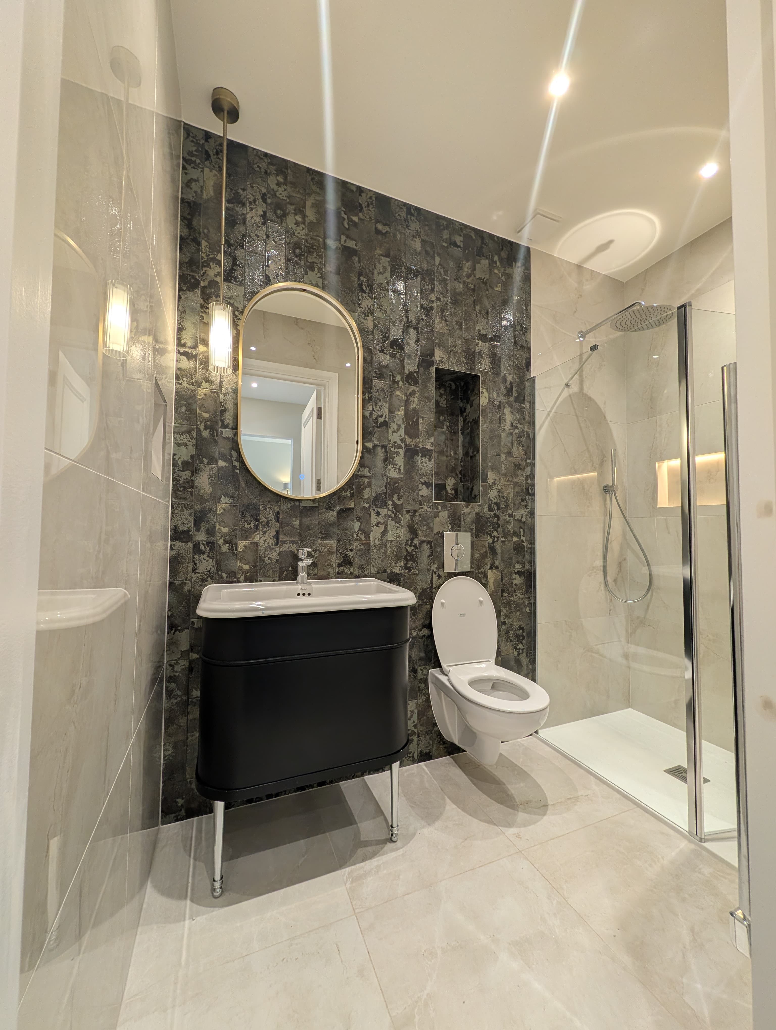 Matt black wall-hung vanity with an oval brass-framed mirror on a dark textured tile feature wall, wall-hung toilet and glass shower screen alongside