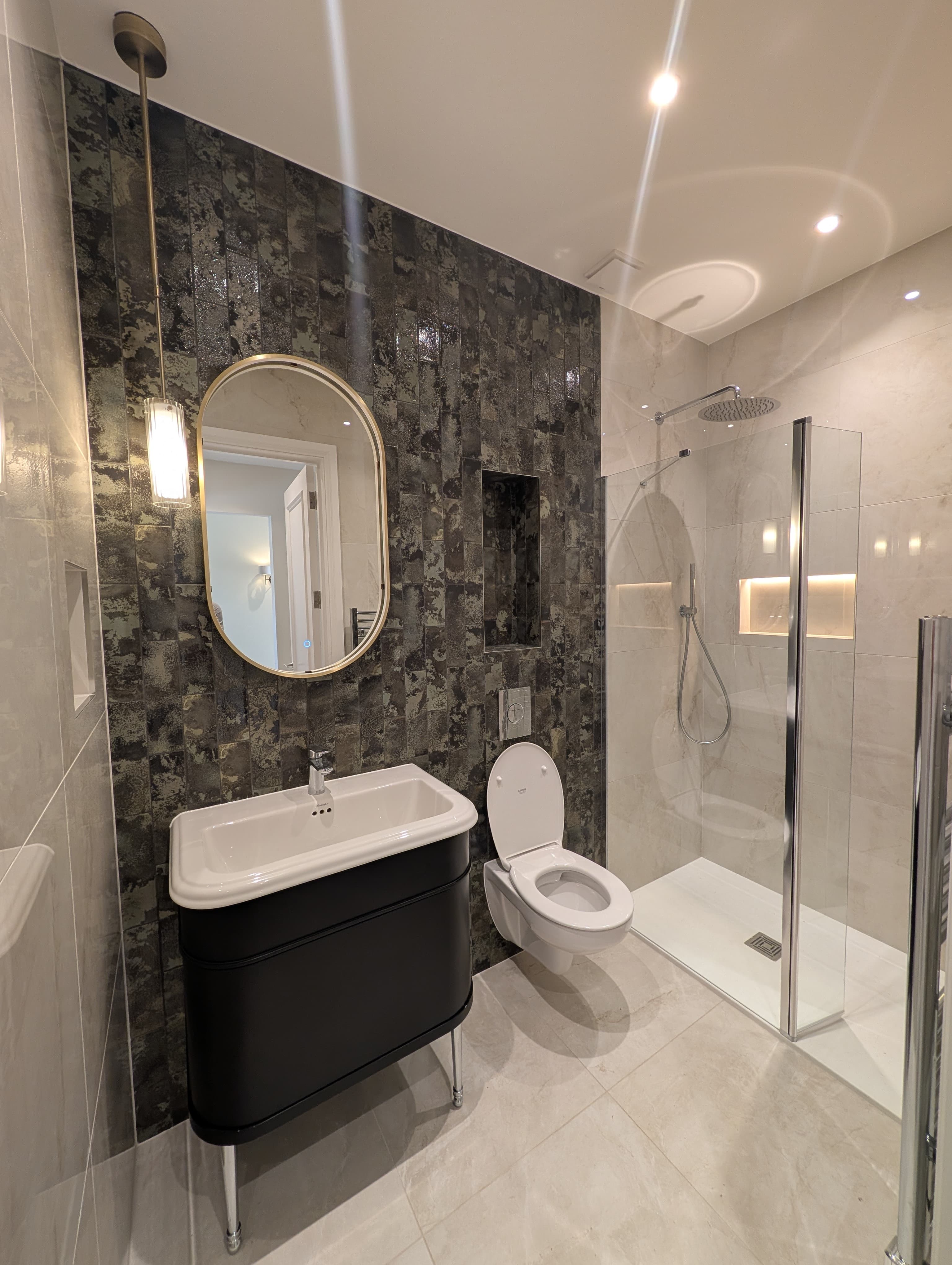 Matt black wall-hung vanity with an oval brass-framed mirror on a dark textured tile feature wall, wall-hung toilet and glass shower screen alongside